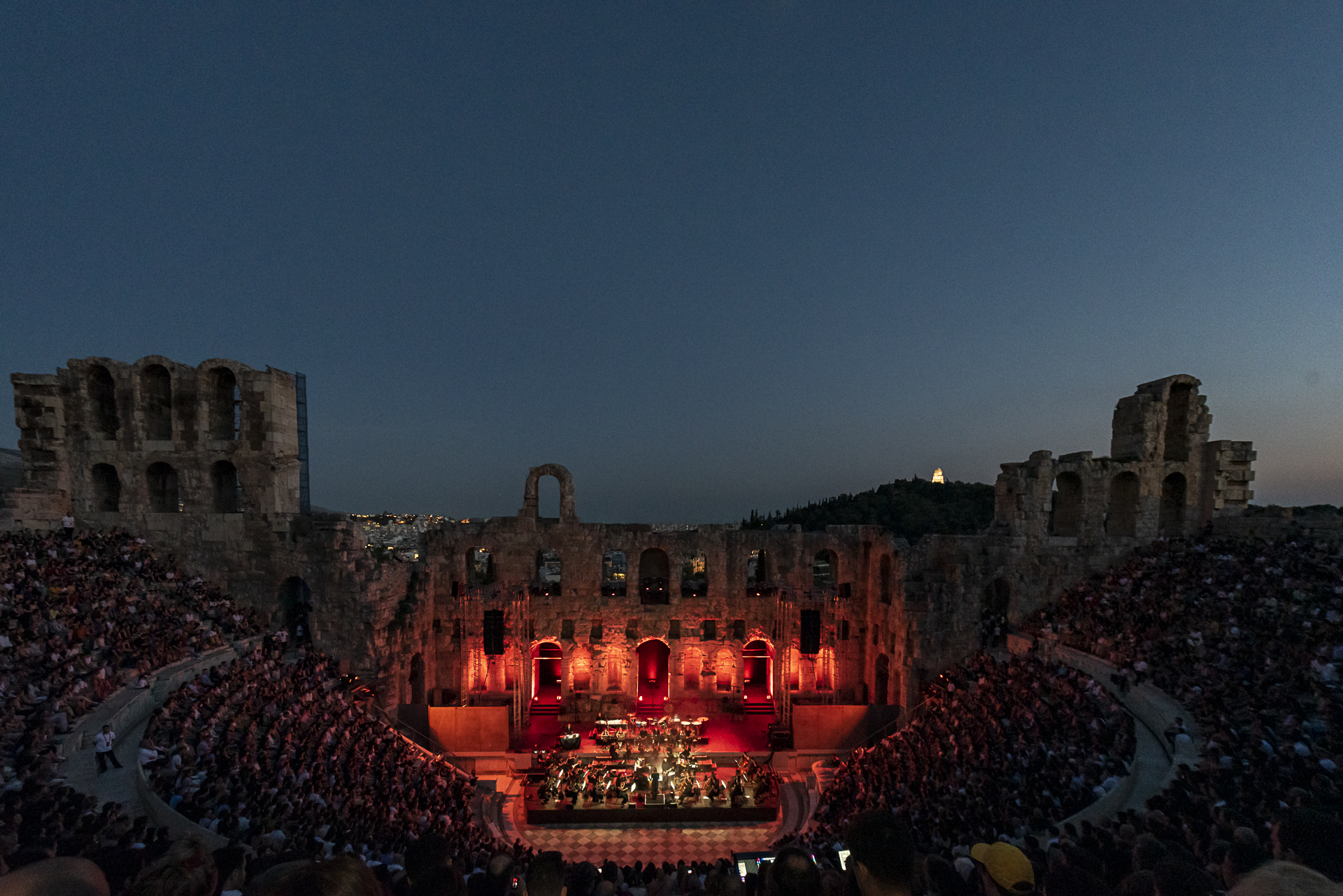 Orchestra playing to a full ancient amphitheatre at dusk, the stage lit deep red