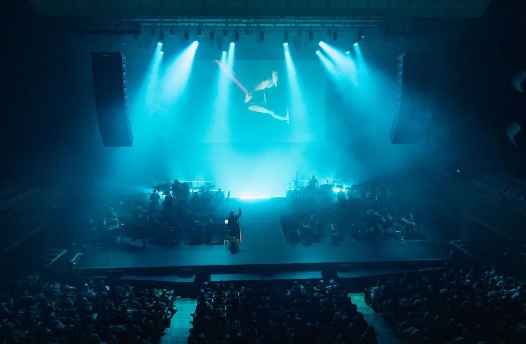 Aerial performer above an orchestra in a hall filled with teal light