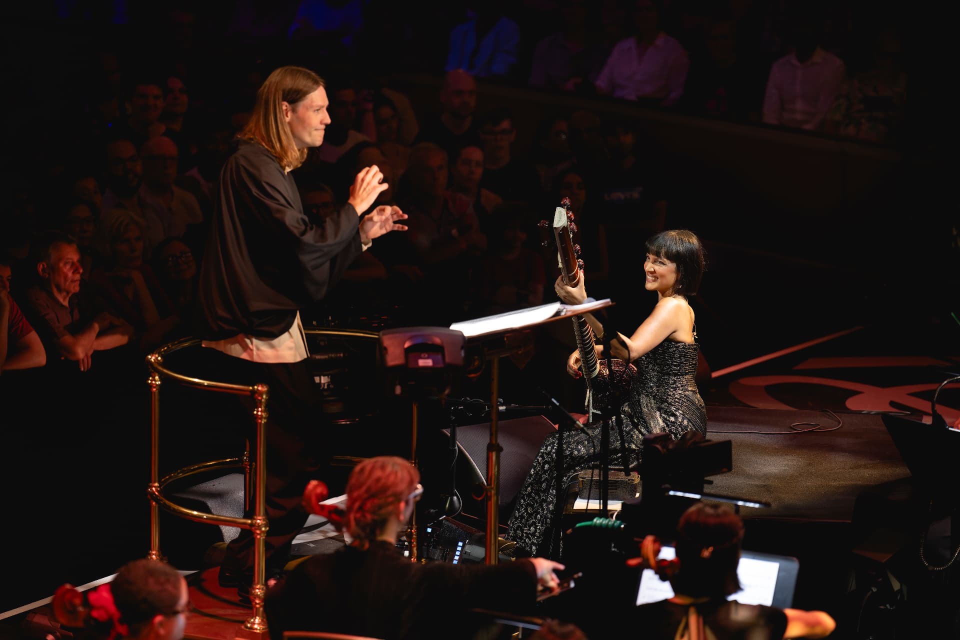 Conductor cueing a smiling soloist holding a stringed instrument on a darkened stage