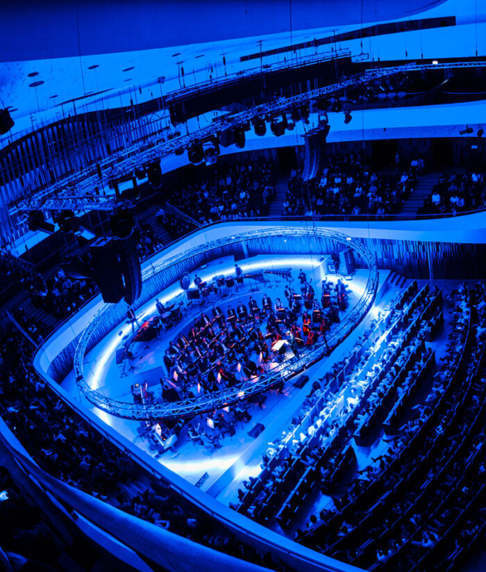 Orchestra in the round inside a modern concert hall flooded with deep blue light
